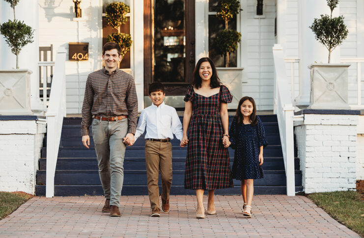 The O'Connor family of four, dressed in casual attire, walks hand in hand on a brick path in front of a white house with blue stairs.