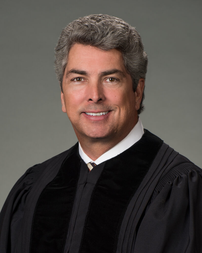 Man in black judge robe with gray hair, smiling against a plain background.