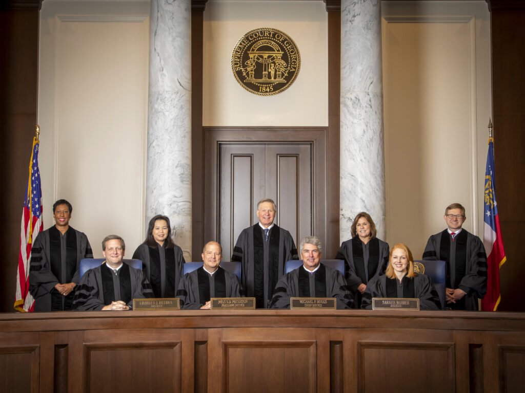 Group of nine judges in robes seated and standing in a courtroom, with flags and a seal in the background.