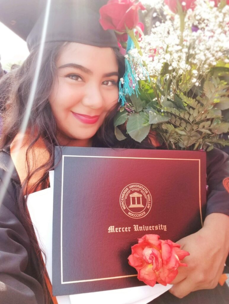 Graduate holding a Mercer University diploma folder and bouquet, wearing a cap and gown, smiling outdoors.