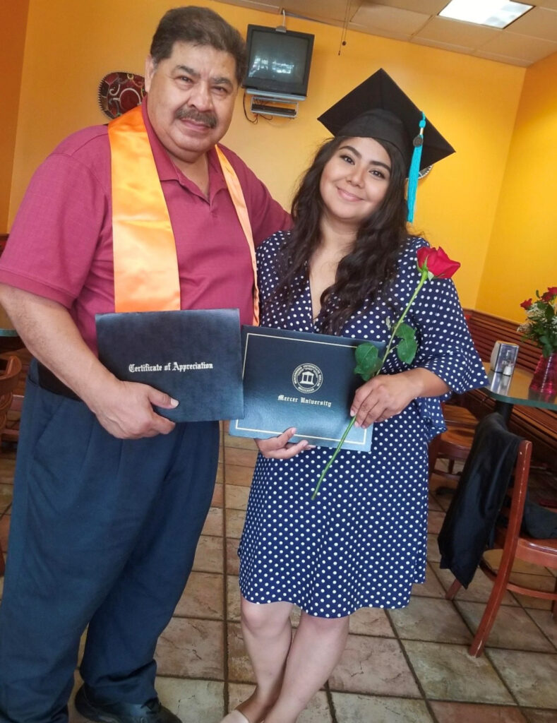 A man and a woman in graduation attire, holding certificates, pose for a photo. The woman holds a red rose.