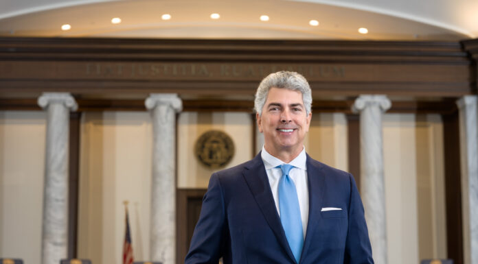 Alumnus, state Supreme Court justice chose law career so he could be a public servant Man in a suit stands confidently in a grand courtroom with columns and a wooden bench in the background.