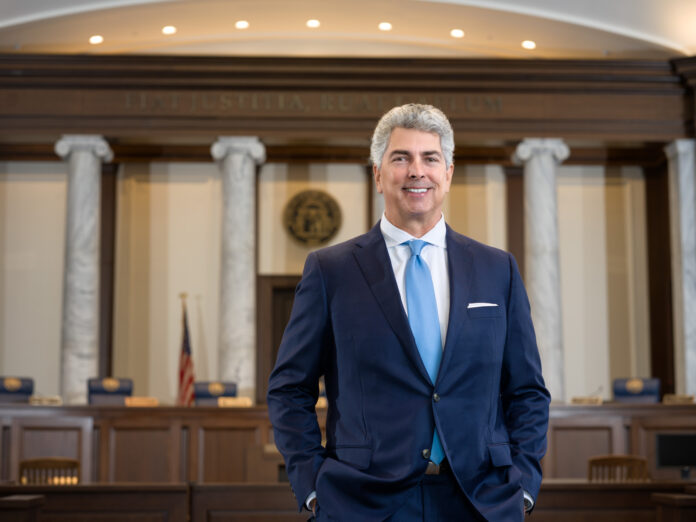 Man in a suit stands confidently in a grand courtroom with columns and a wooden bench in the background.