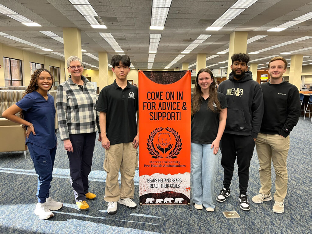 Five students and a professor stand in a library next to a sign for Mercer University Pre-Health Ambassadors.