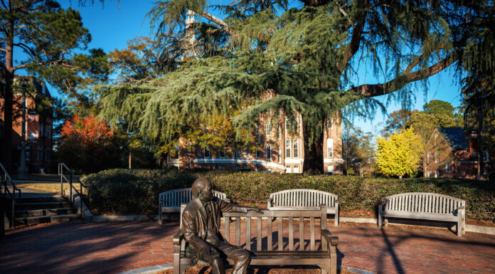 Faculty and Staff Notables | March 2025 Bronze statue of a person sitting on a bench in a park-like setting, surrounded by trees and buildings under a blue sky.