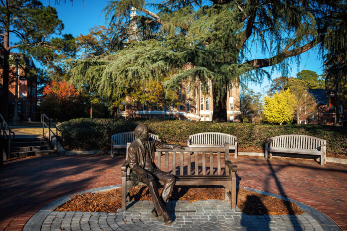 Bronze statue of a person sitting on a bench in a park-like setting, surrounded by trees and buildings under a blue sky.