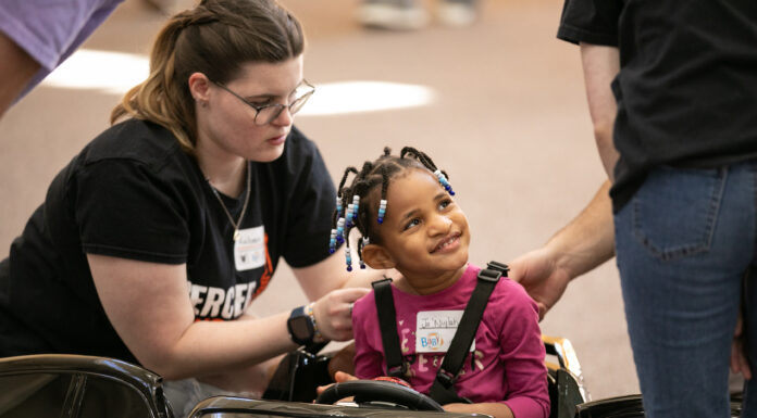 Go Baby Go! Students to modify toy cars for kids with limited mobility Child in a toy car is helped by two adults. The child is smiling and wearing a pink shirt and name tag, while one adult adjusts the seatbelt.