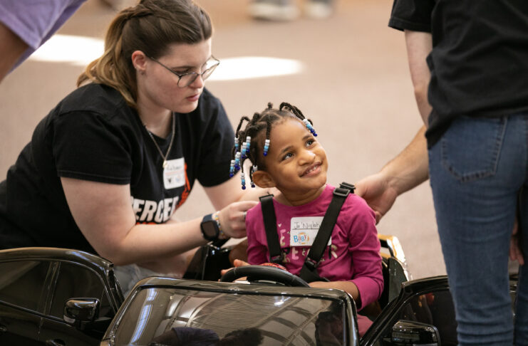 Child in a toy car is helped by two adults. The child is smiling and wearing a pink shirt and name tag, while one adult adjusts the seatbelt.