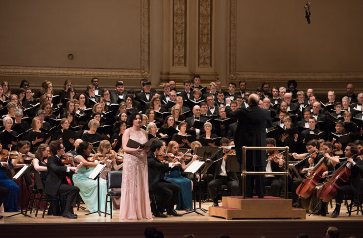 An orchestra and choir perform on stage, with a soloist standing in front of the musicians and conductor.