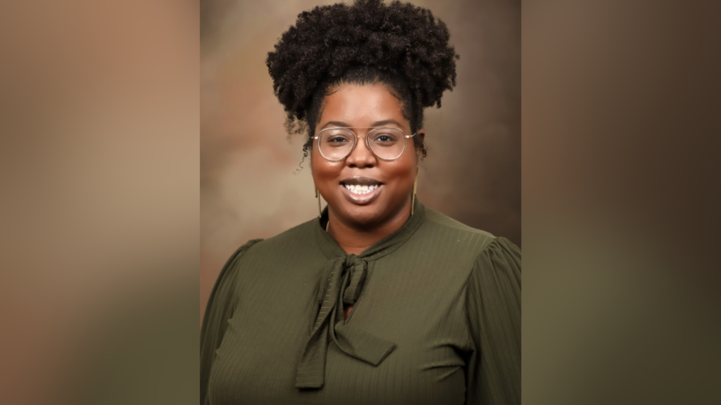 Person wearing glasses and an olive green top, smiling against a brown background.
