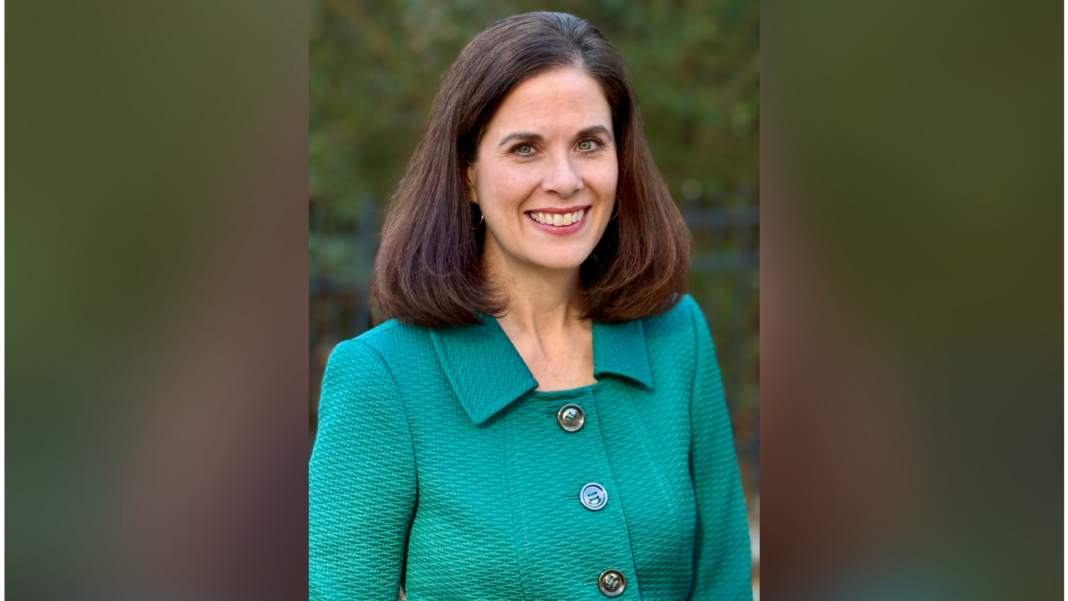 Woman with brown hair smiling, wearing a green textured jacket, standing outside with greenery in the background.