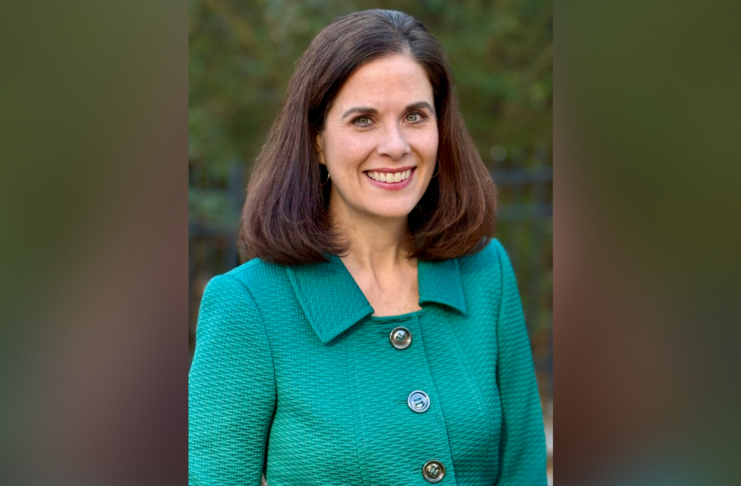 Woman with brown hair smiling, wearing a green textured jacket, standing outside with greenery in the background.