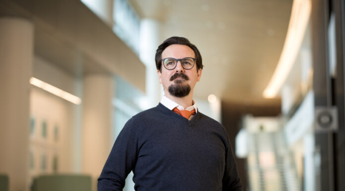 Leadership experiences at Mercer shaped direction of alumnus’s nursing career Man with glasses and a beard stands in a modern, well-lit building, wearing a dark sweater, white shirt, and orange tie.