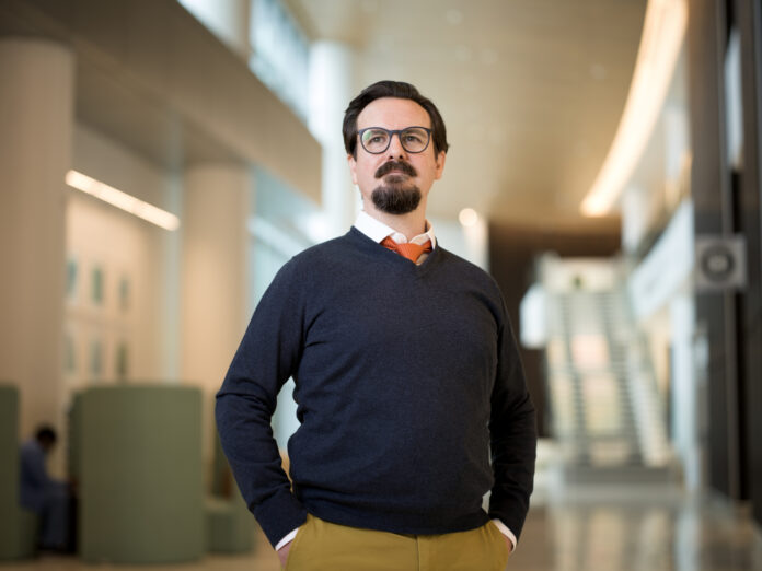 Man with glasses and a beard stands in a modern, well-lit building, wearing a dark sweater, white shirt, and orange tie.