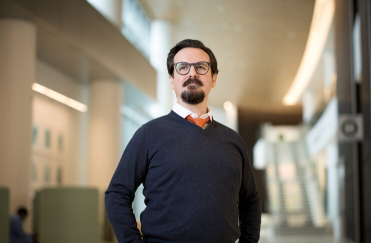 Man with glasses and a beard stands in a modern, well-lit building, wearing a dark sweater, white shirt, and orange tie.