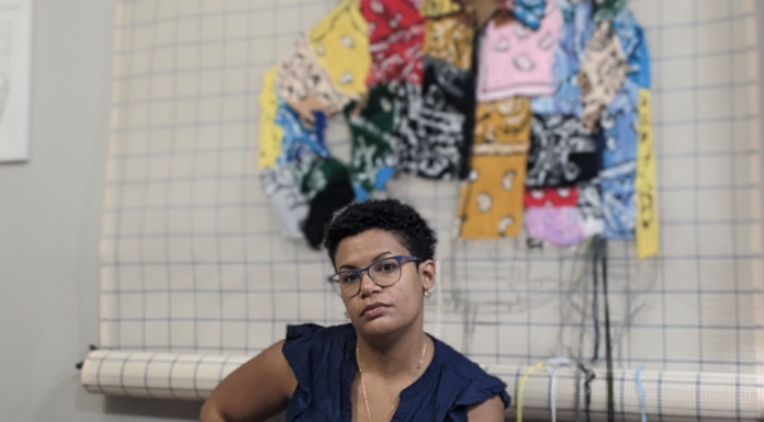 McEachern Art Center presents Afro-Caribbean figurative artist Kandy Lopez in “Vaivén” Person sitting on a stool in front of woven textile art. They are wearing glasses and a blue shirt.