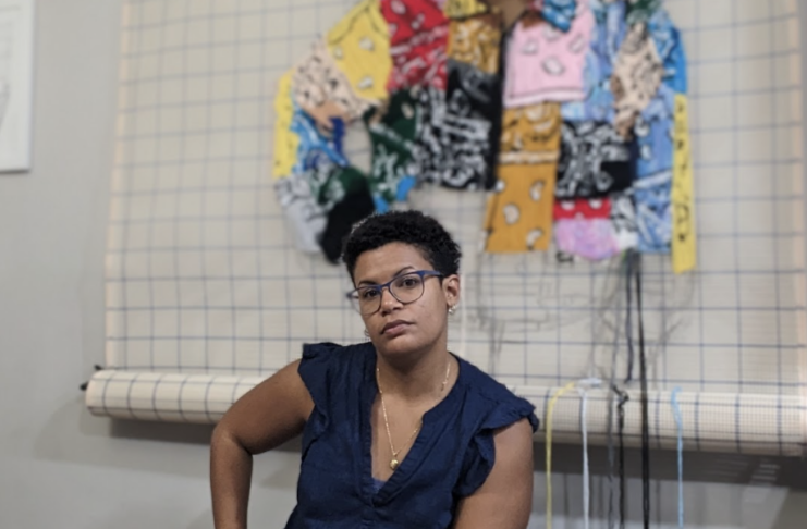 Person sitting on a stool in front of woven textile art. They are wearing glasses and a blue shirt.