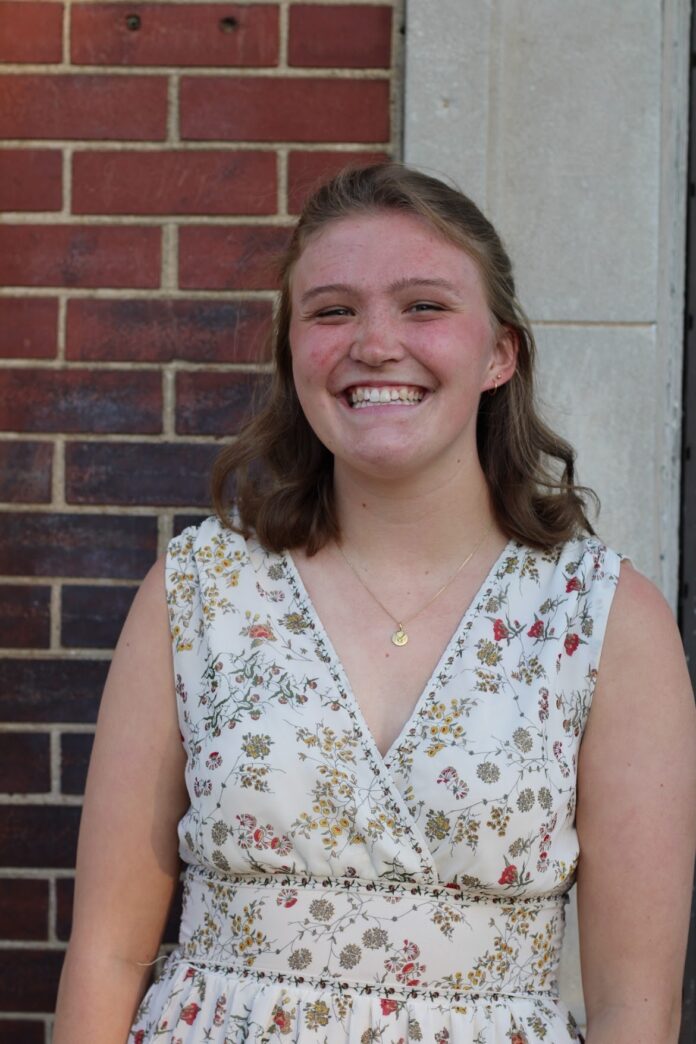 Person smiling, standing in front of a brick and stone wall, wearing a floral dress.