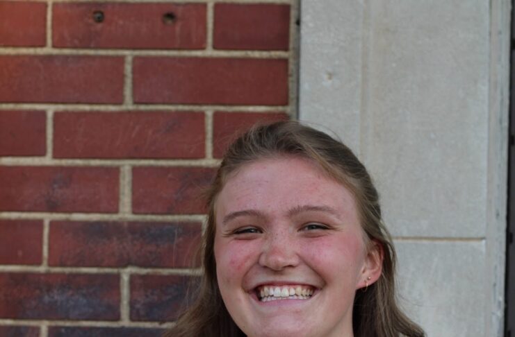 Person smiling, standing in front of a brick and stone wall, wearing a floral dress.