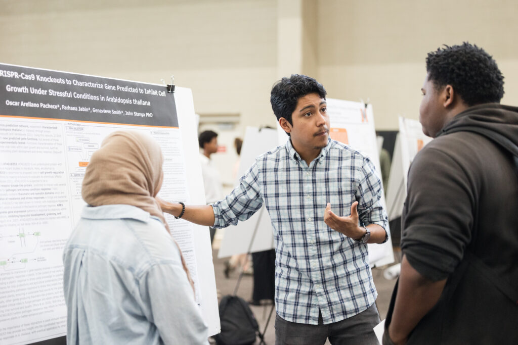 A man presents research during a poster session, discussing with two attendees in a conference setting.