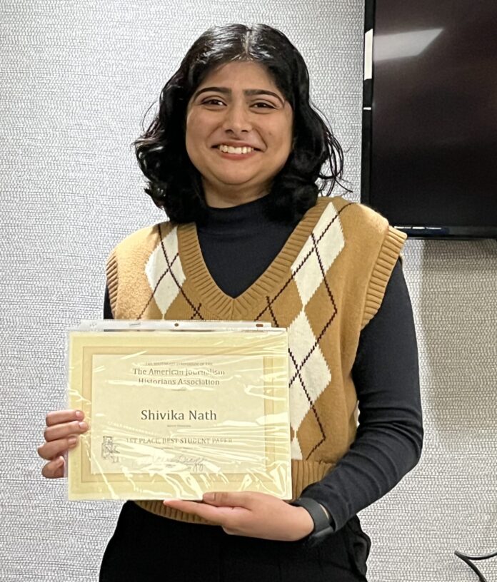 Person holding a certificate and smiling, wearing a patterned brown vest over a black shirt, in front of a textured wall.