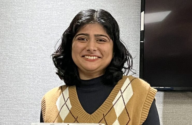 Person holding a certificate and smiling, wearing a patterned brown vest over a black shirt, in front of a textured wall.