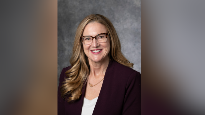 Woman with long hair in a purple blazer and glasses, smiling against a gray background.