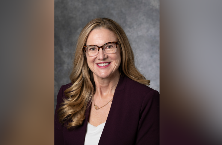 Woman with long hair in a purple blazer and glasses, smiling against a gray background.