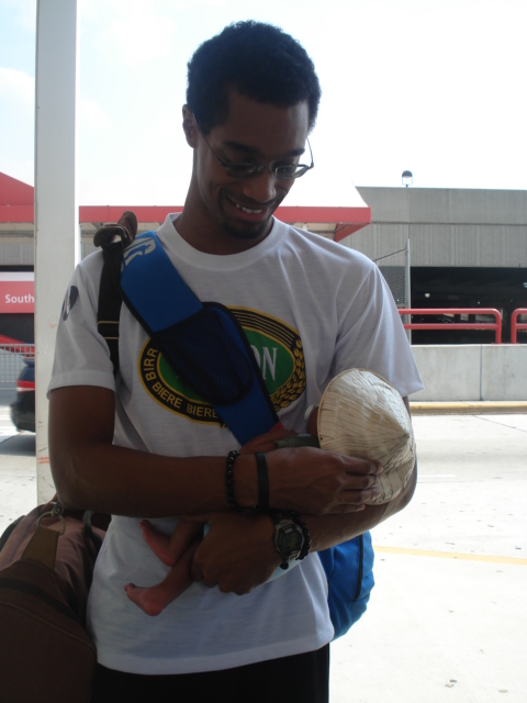 A man wearing glasses and a patterned T-shirt smiles while holding a sleeping baby wrapped in a light-colored blanket.