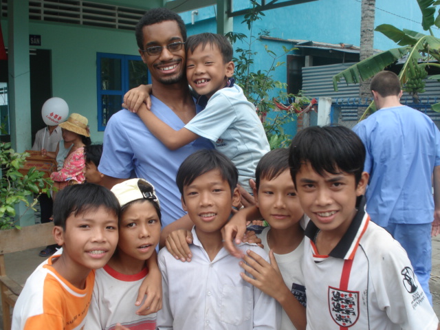 A man in scrubs smiles, surrounded by six young boys, posing outdoors in a casual setting with a building in the background.