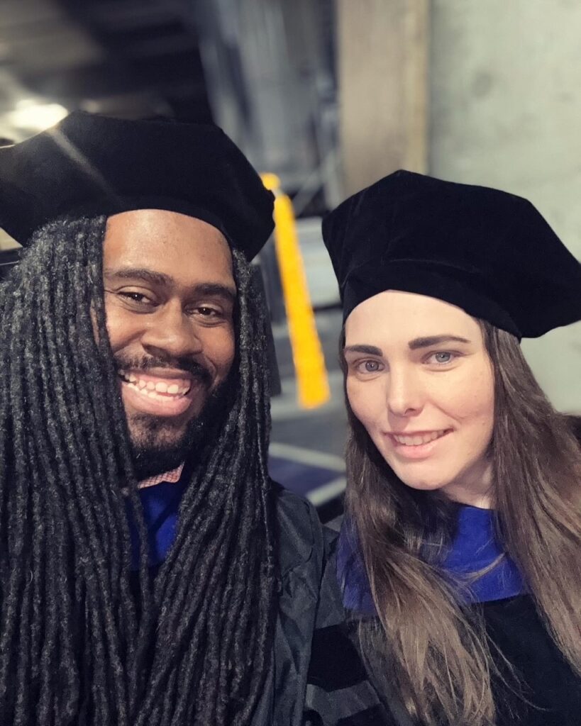 Two people wearing graduation regalia, including black caps and gowns, smiling for a photo indoors.