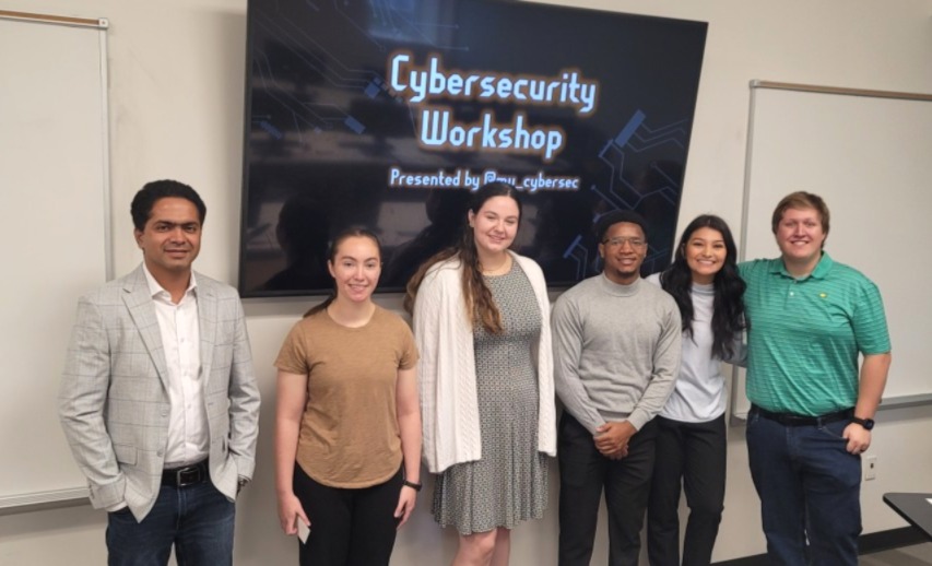 Six people standing in front of a screen displaying Cybersecurity Workshop Presented by BWI_cybersec.