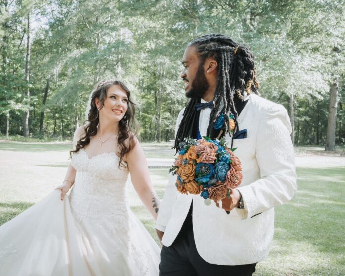 Bride and groom walking outside, smiling at each other; the groom holds a bouquet.