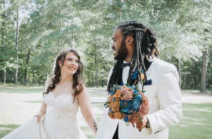 Bride and groom walking outside, smiling at each other; the groom holds a bouquet.