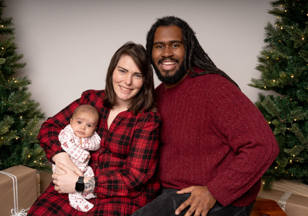 A couple sits between decorated Christmas trees, holding a baby in festive pajamas, with a wrapped gift nearby.