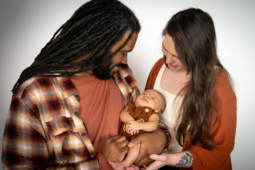 A couple holds a sleeping baby between them. The man has dreadlocks and wears a plaid shirt; the woman has long hair and a tattoo.