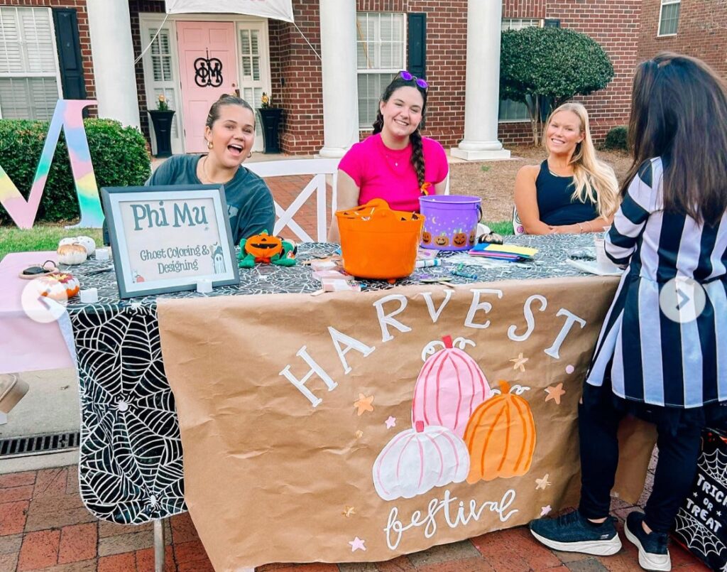 Three women sit behind a decorated table labeled Harvest Festival outside a brick building, interacting with a standing person.