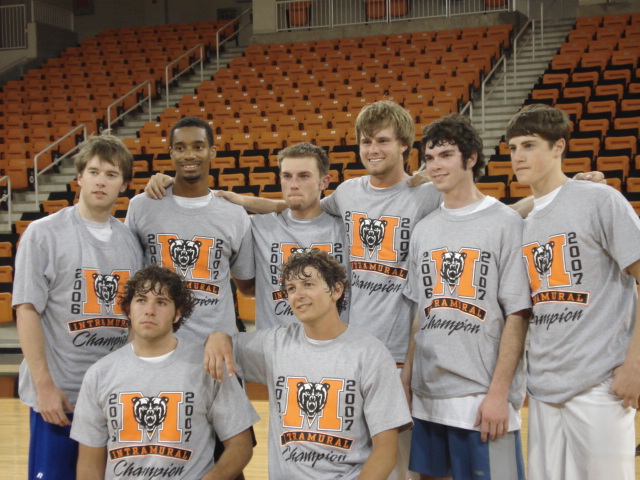 Eight young men wearing matching shirts pose together on a basketball court with empty stands in the background.