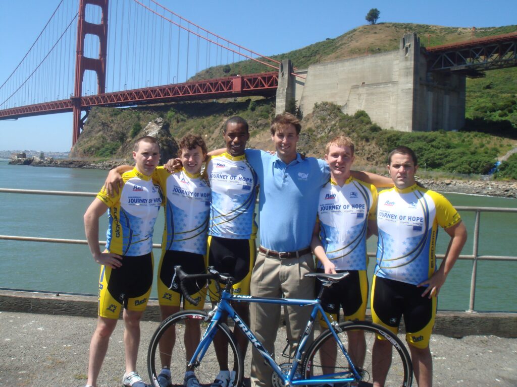 Six people in cycling jerseys pose with a bike in front of the Golden Gate Bridge.