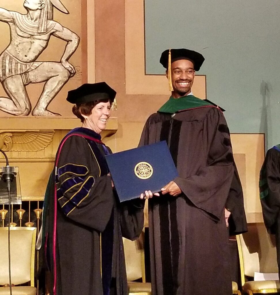 A graduate in a cap and gown receives a diploma from a faculty member during a ceremony, with others seated and standing nearby.