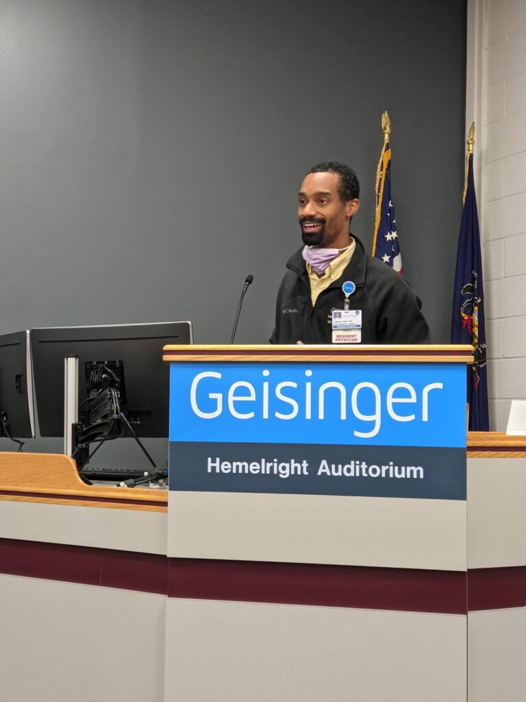 A person stands at a podium labeled Geisinger Hemelright Auditorium, with computers and flags in the background.