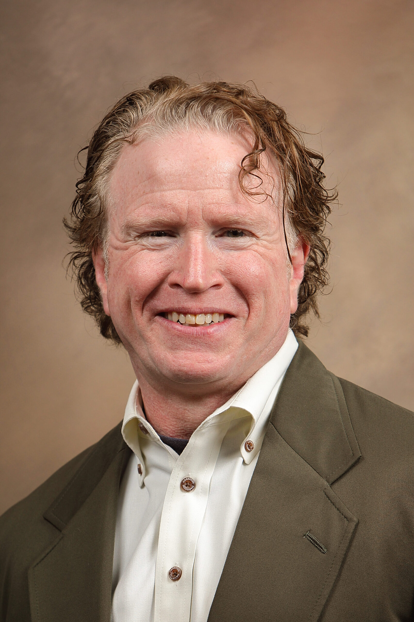 Man with curly hair wearing a white shirt and olive green blazer, smiling against a brown background.