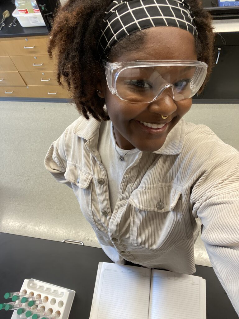 Young woman wearing safety goggles smiling in a lab. She is standing by an open notebook and test tubes.