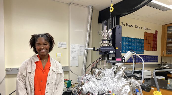 Junior discovers interest in pharmacy and research at Mercer Asia Loney stands in a lab next to scientific equipment covered in foil, with a periodic table visible on the wall behind.