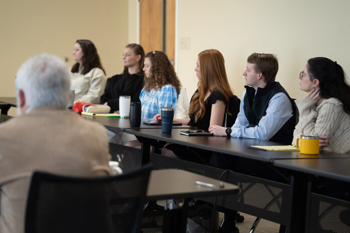 A group of people seated at desks in a classroom, attentively listening to someone out of view.