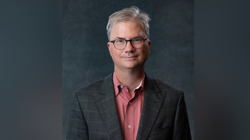 Man with glasses in a plaid jacket and red shirt standing against a dark gray background.