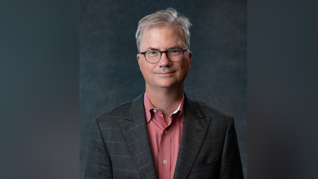 Man with glasses in a plaid jacket and red shirt standing against a dark gray background.