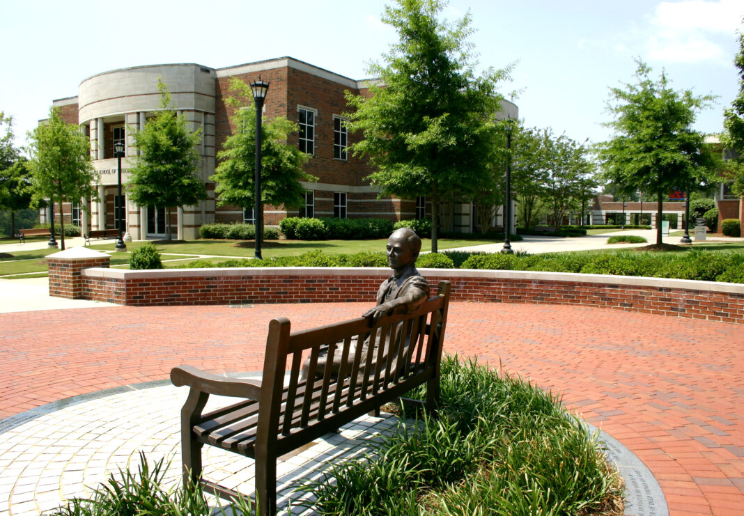 Bronze sculpture of a person sitting on a bench in front of a brick building with circular architecture and green landscape.