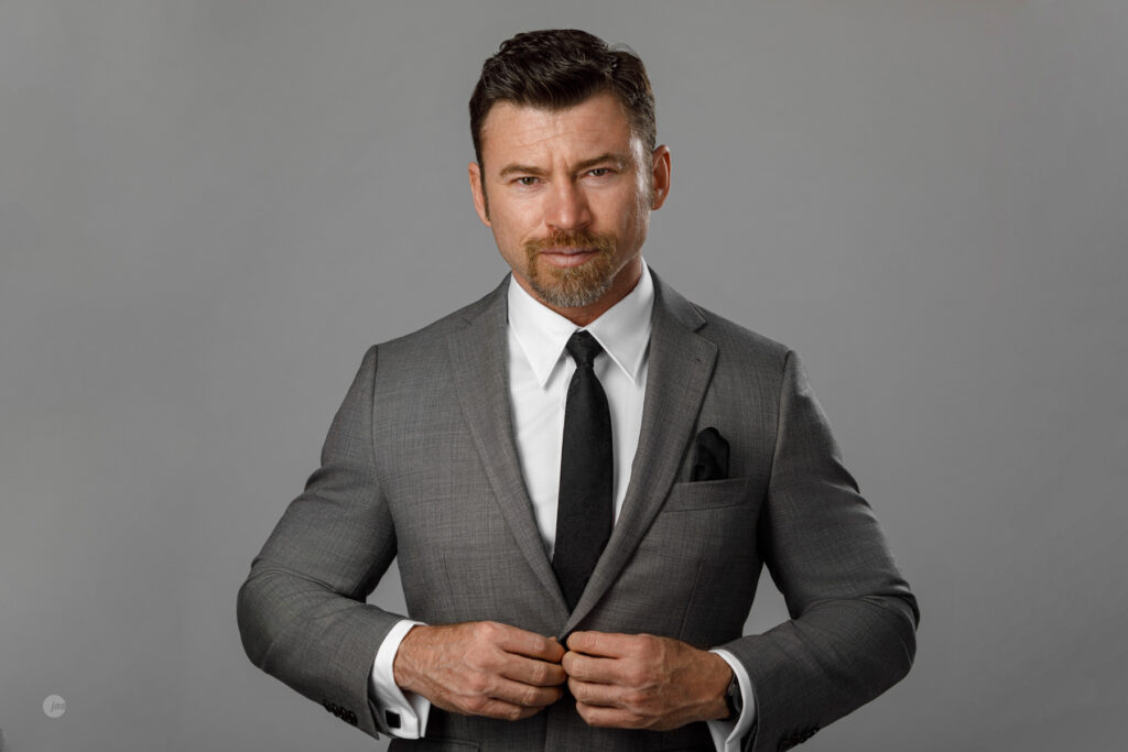 A man in a gray suit with a black tie and pocket square stands against a gray background, buttoning his jacket.