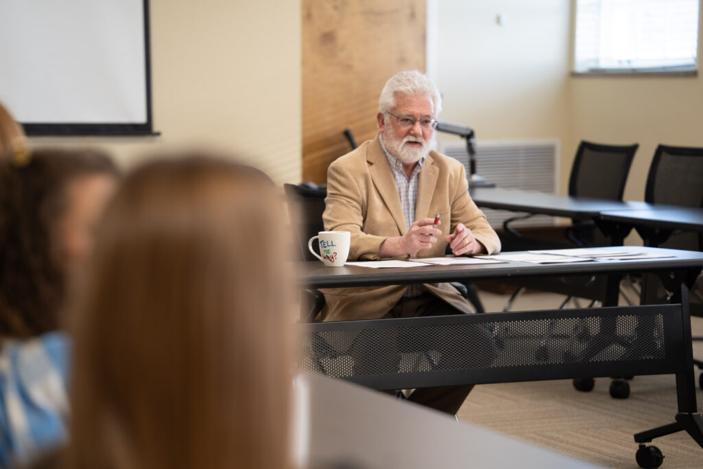 Pat Longan sits at a table, holding a pen and gesturing.